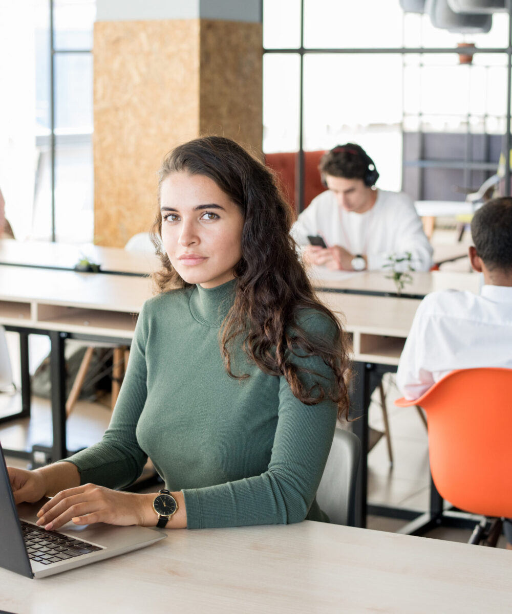 Portrait of beautiful young woman looking at camera while working with laptop in modem open office, copy space