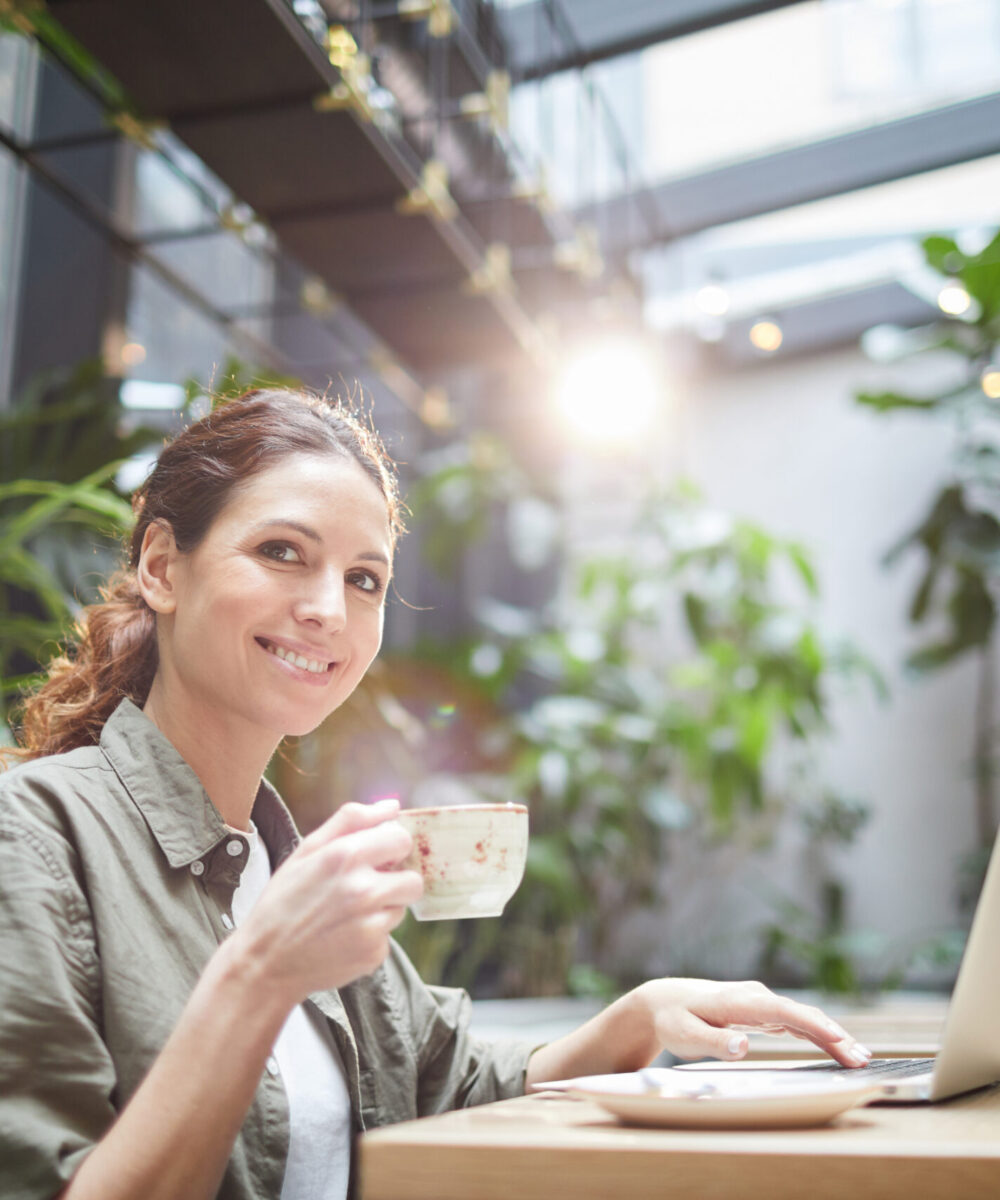 Portrait of smiling young woman looking at camera while working with laptop in cafe and enjoying coffee, copy space